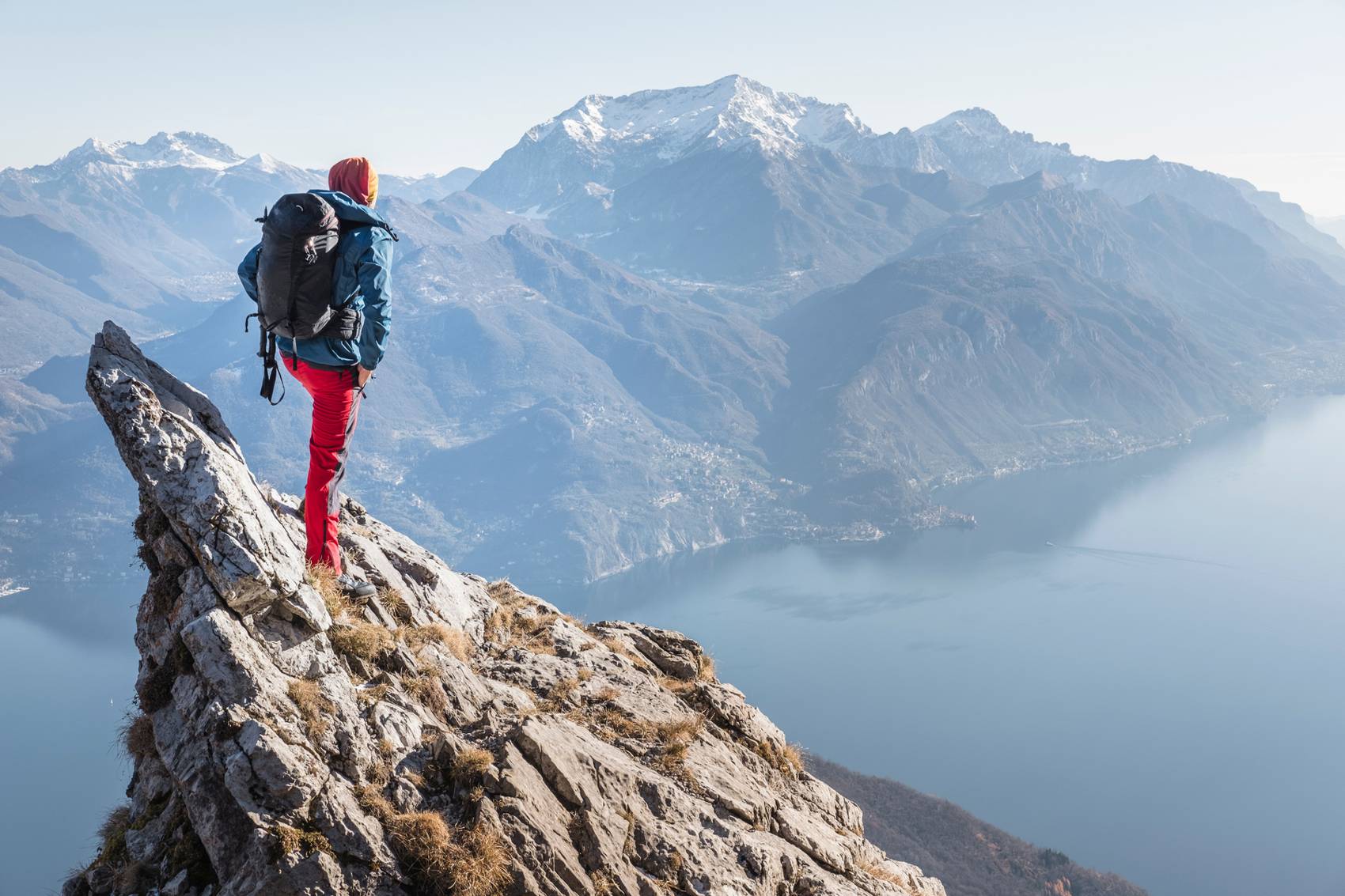 A hiker in a red jacket stands on a rocky ridge above a mountain lake